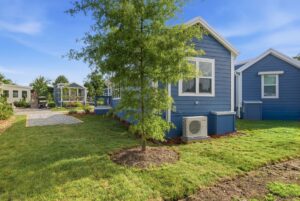A blue house with white trim sits on a grassy yard with a tree, gravel driveway, and outdoor AC unit.