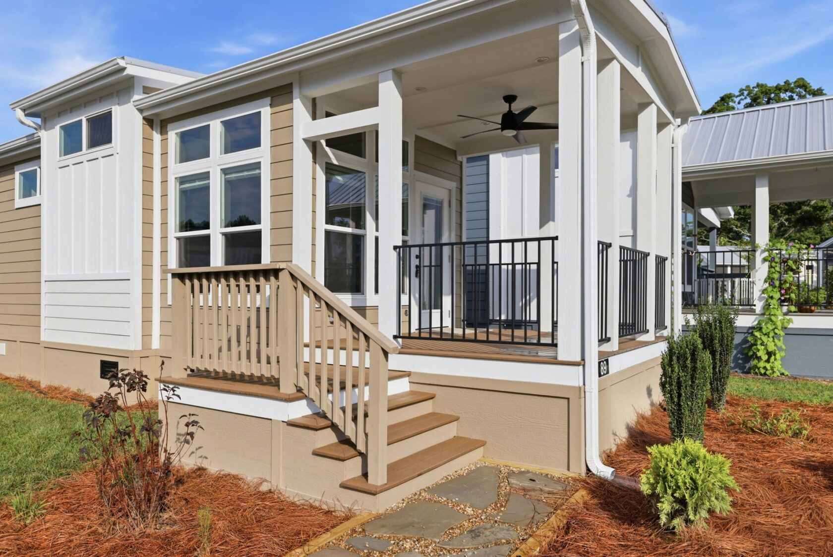 Beige house with white trim, a porch with black railing, steps, mulch, and a stone walkway in front.