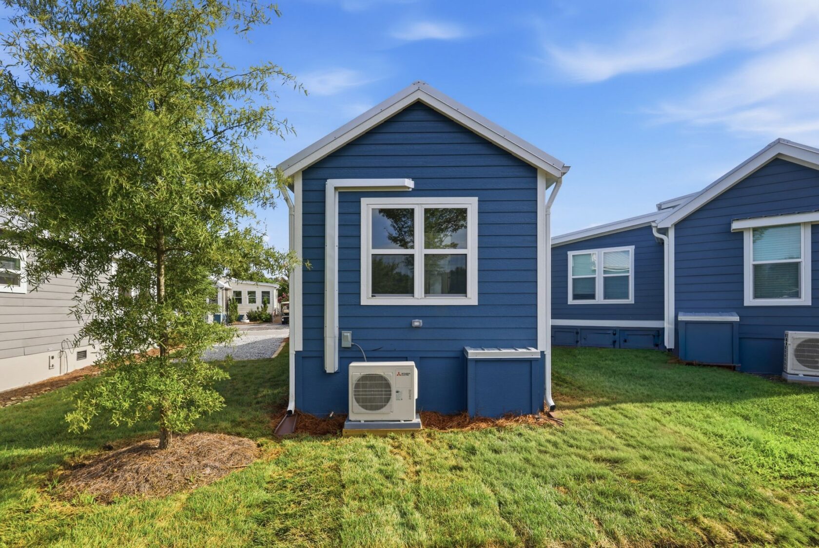 Small blue house with white trim, an air conditioning unit, and a tree on a green lawn under a blue sky.
