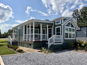 Green tiny house with white trim and large windows sits on Meandering Lane, wraparound porch beneath a blue sky.
