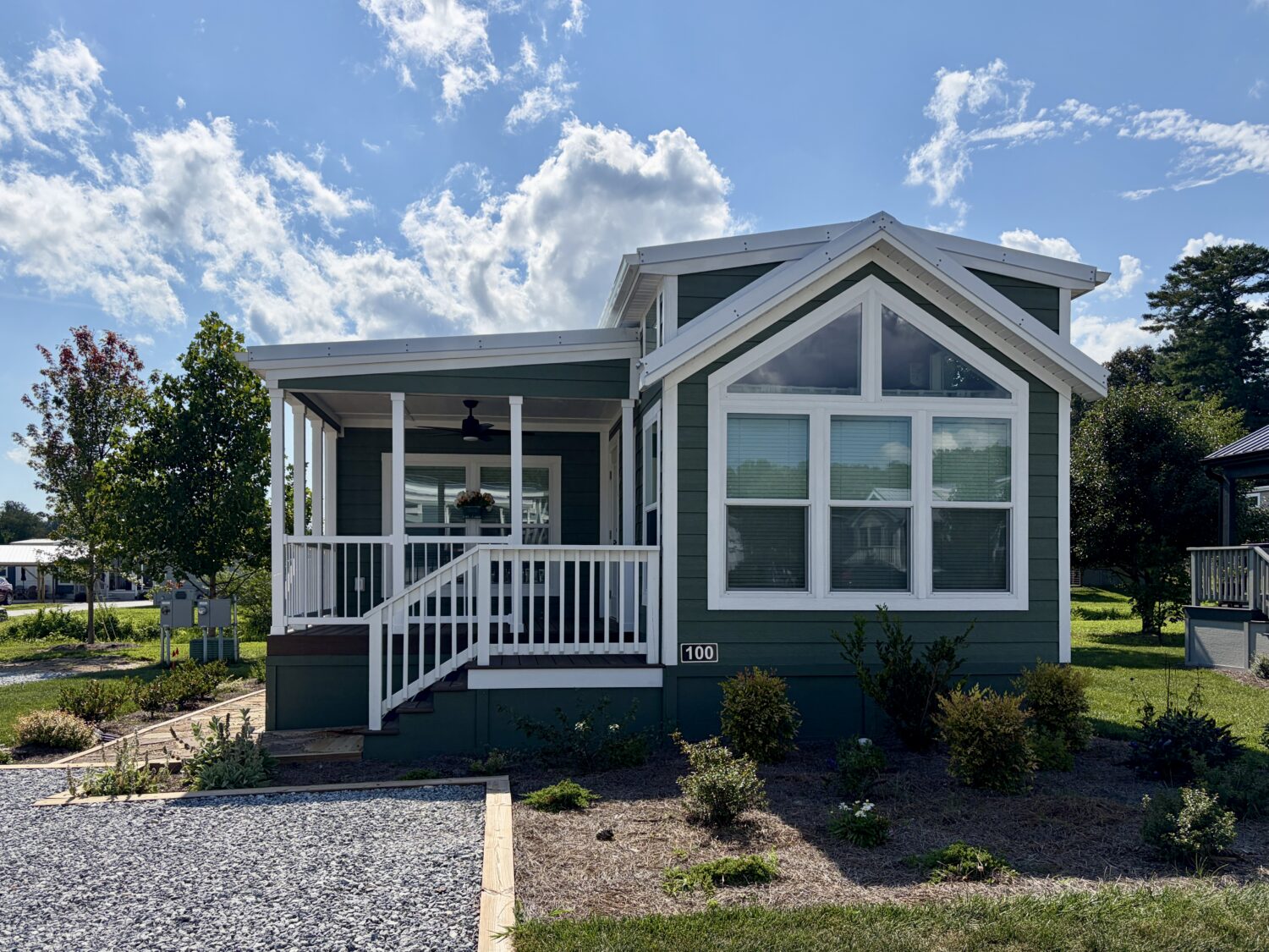 Green manufactured home at 100 Meandering Lane features large windows, white trim, a porch, and a landscaped yard.