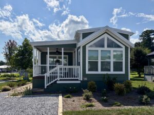 Green manufactured home at 100 Meandering Lane features large windows, white trim, a porch, and a landscaped yard.