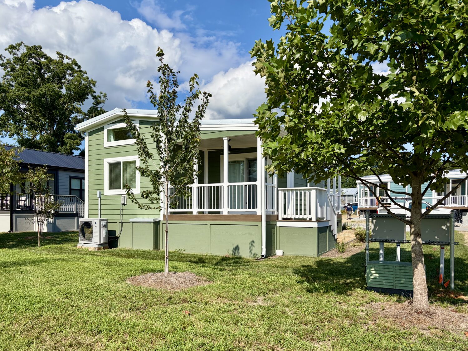 A small green house with a porch on Meandering Lane, trees, and grass yard under a partly cloudy blue sky.