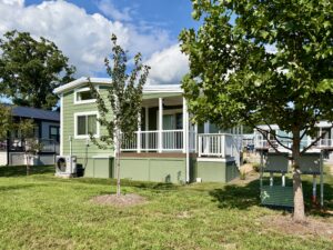 A small green house with a porch on Meandering Lane, trees, and grass yard under a partly cloudy blue sky.