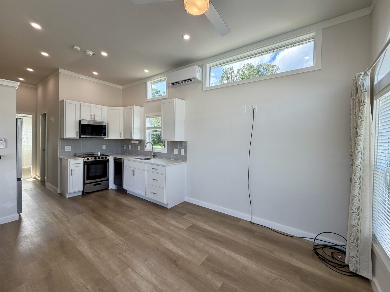Modern kitchen on Meandering Lane features white cabinets, wood floors, high windows, and an empty living area.