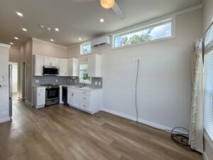 Modern kitchen on Meandering Lane features white cabinets, wood floors, high windows, and an empty living area.