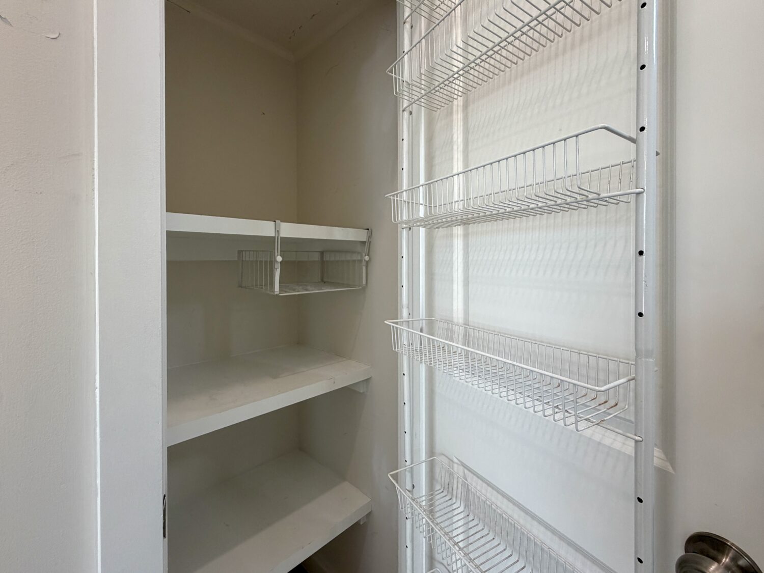 Empty pantry at 100 Meandering Lane with white shelves and metal wire baskets on the door, viewed from the entrance.