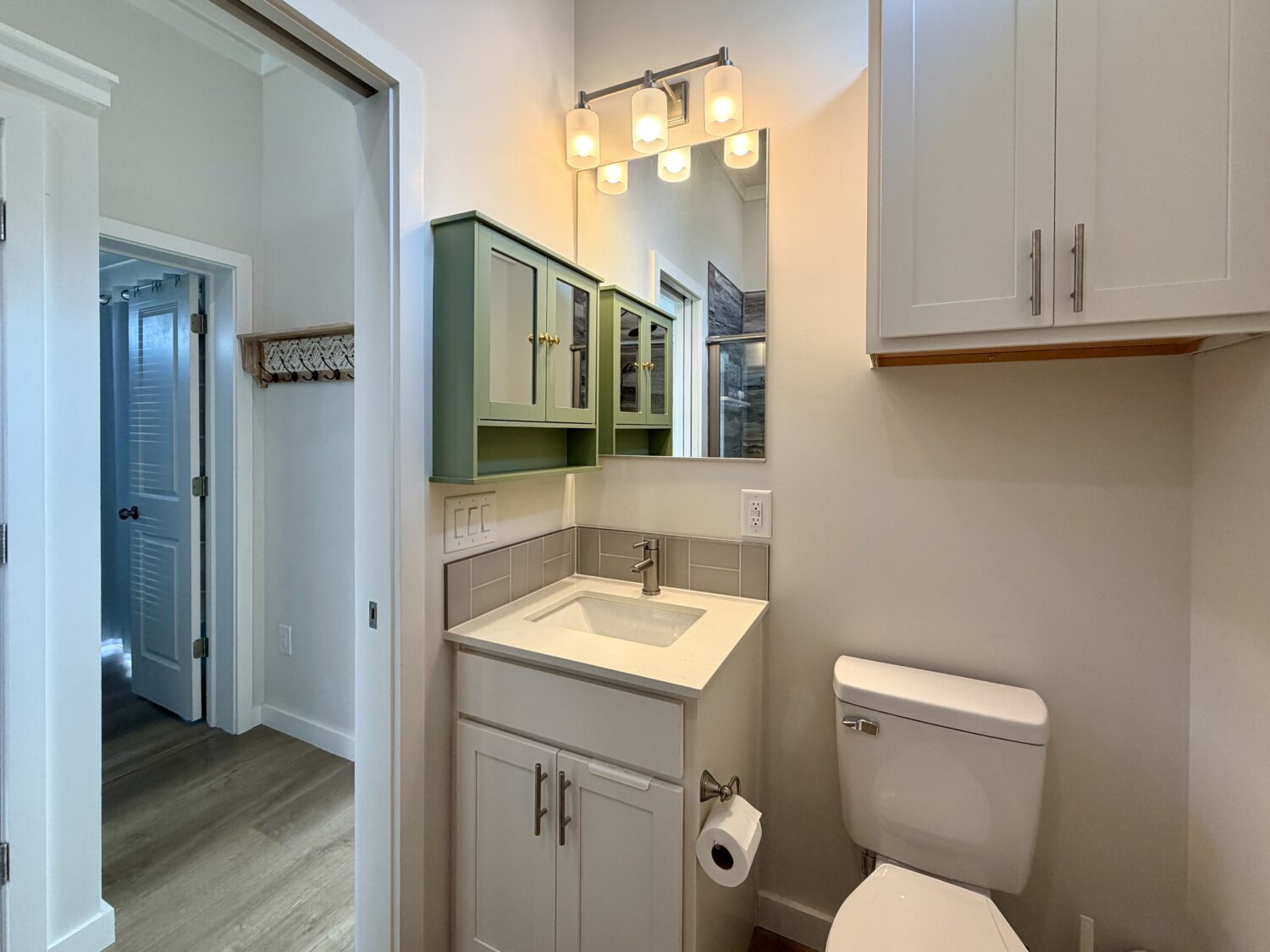 Bathroom with sink, green and white cabinets, mirror, toilet, and doorway to another room on Meandering Lane.