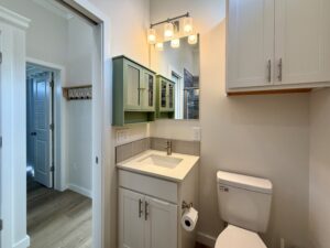 Bathroom with sink, green and white cabinets, mirror, toilet, and doorway to another room on Meandering Lane.