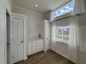 White bedroom on Meandering Lane with wood floor, dresser, two closets, and sunny window with sheer curtains.
