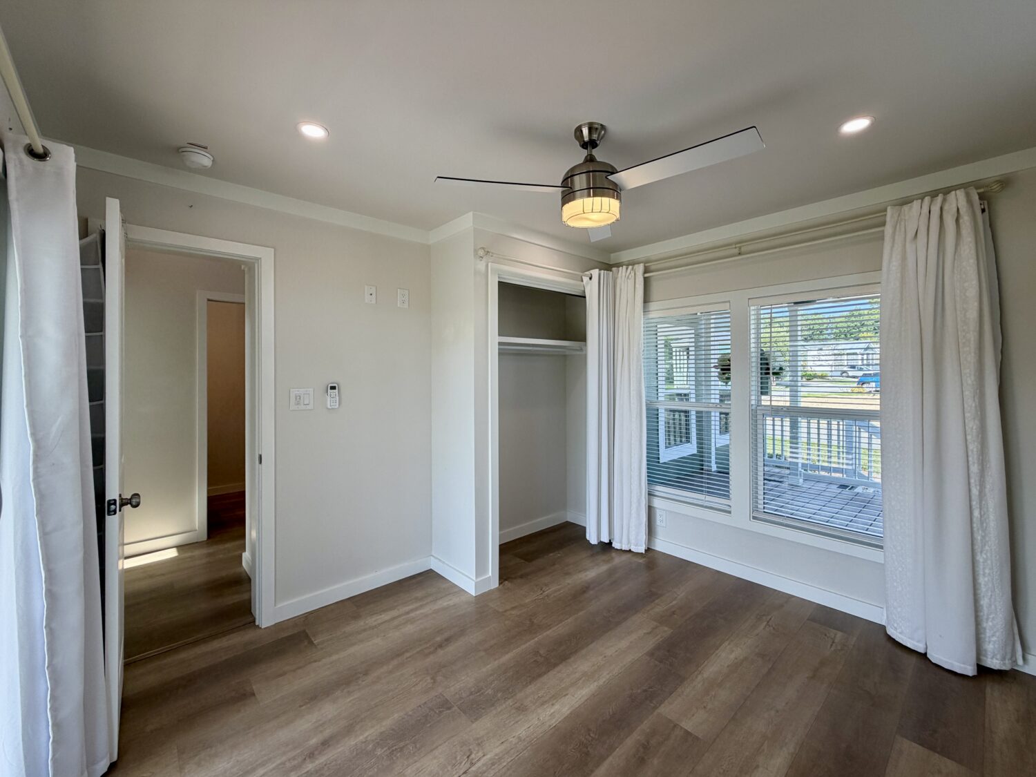 A bright, empty room at 100 Meandering Lane with wood floors, ceiling fan, closet, and large windows with white curtains.