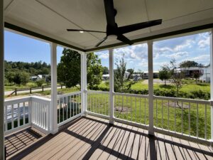 A covered porch on Meandering Lane with a ceiling fan overlooks a green yard, white railing, and sunny neighborhood views.