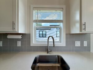 Modern kitchen sink with faucet, gray tile backsplash, and a window overlooking Meandering Lane and a charming house.