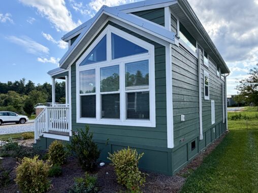 Green tiny house with white trim on Meandering Lane, large windows, a small porch, and lush greenery all around.