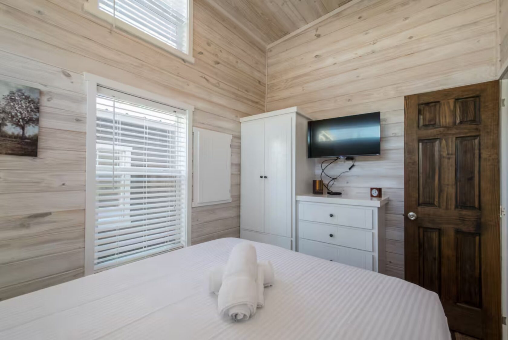 Small bedroom at 66 Empire Lane with light wood walls, a bed, white dresser, TV, wardrobe, and window with blinds.