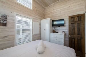 Small bedroom at 66 Empire Lane with light wood walls, a bed, white dresser, TV, wardrobe, and window with blinds.