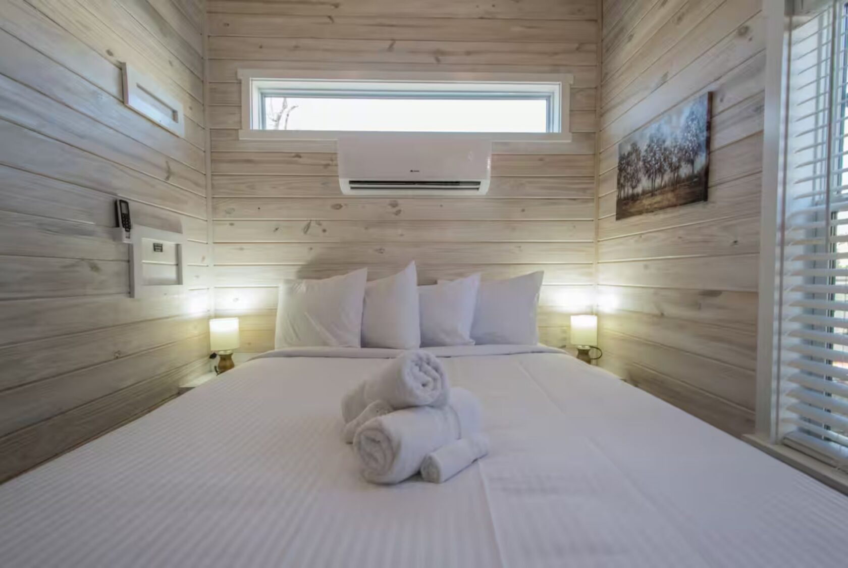 A neatly made bed with white linens and folded towels in a wood-paneled room at 66 Empire Lane, with window and wall art.