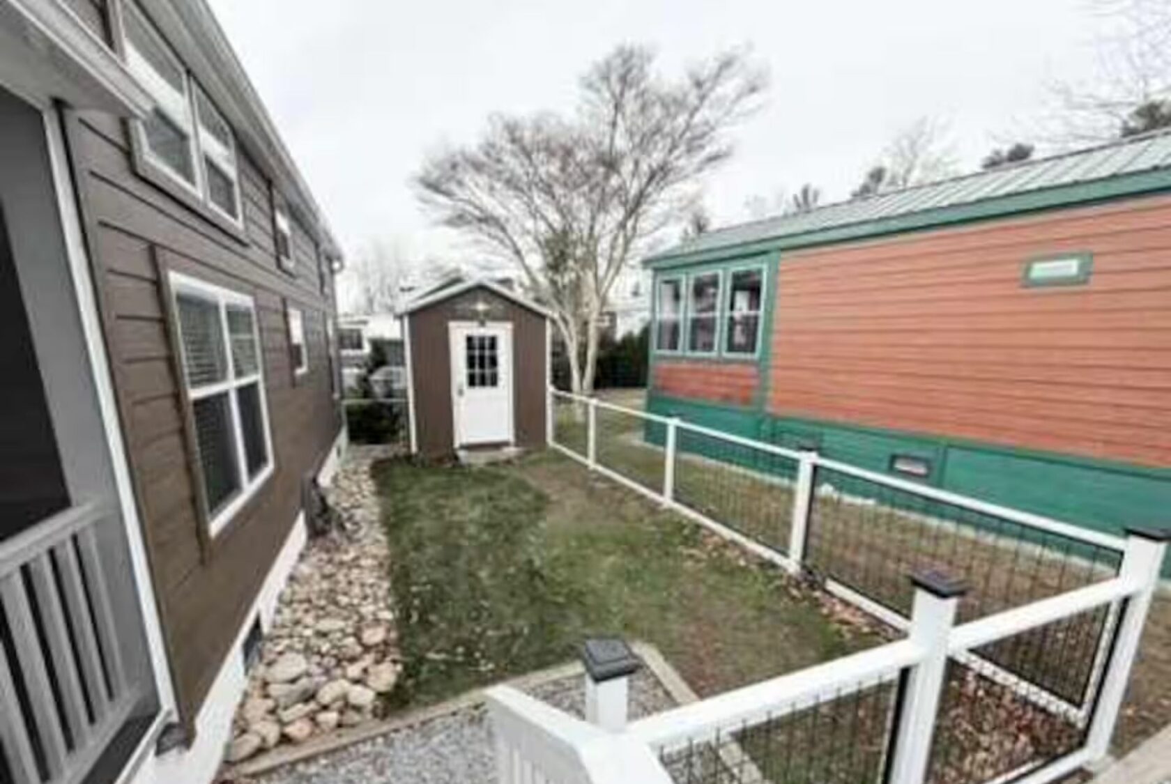 A fenced yard at 66 Empire Lane with a small shed between two tiny houses. Rocks line one side on a cloudy day.