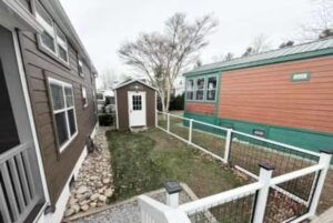 A fenced yard at 66 Empire Lane with a small shed between two tiny houses. Rocks line one side on a cloudy day.