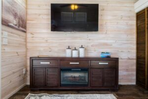 A wall-mounted TV above a dark wood console at 66 Empire Lane, with books, jars, and an electric fireplace.