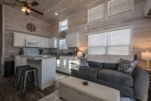 Cozy modern kitchen and living room at 66 Empire Lane with gray sofa, white cabinets, bar stools, and window blinds.