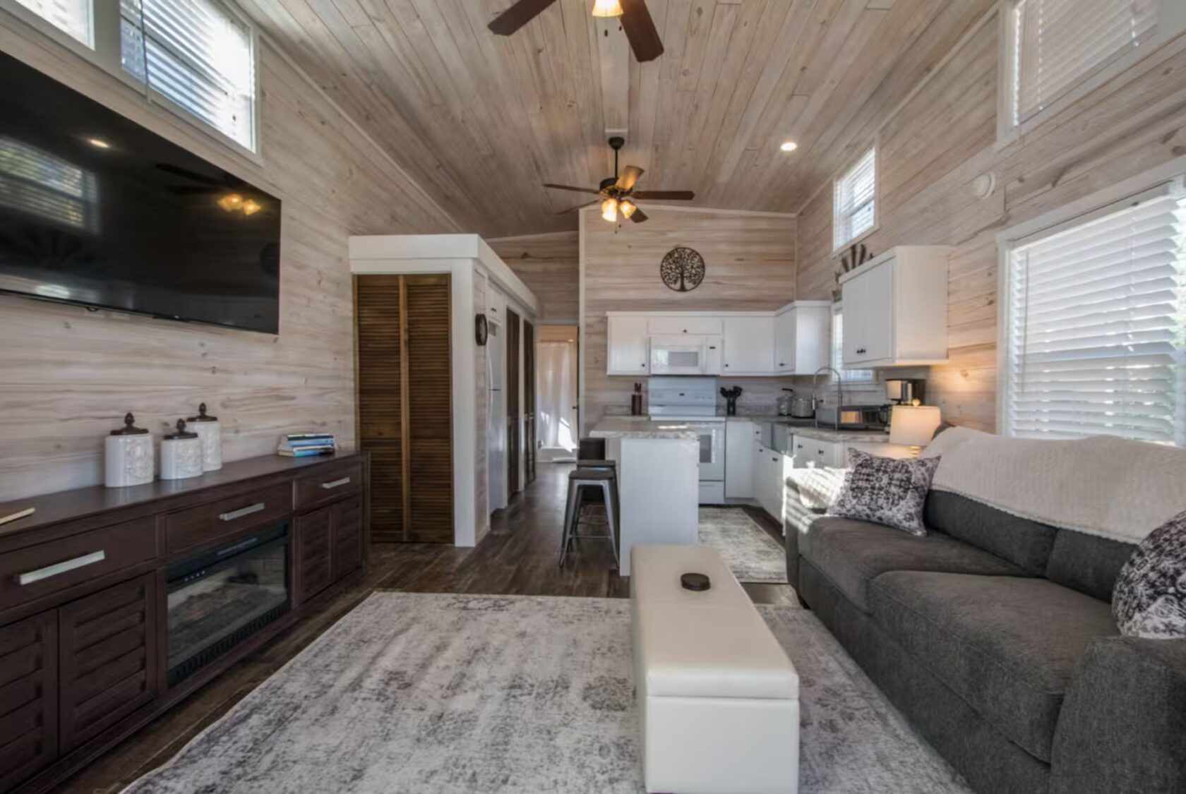 Modern living room at 66 Empire Lane with wood paneling, gray sofa, white kitchen, island, TV, and ceiling fan.